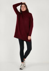 Bordeauxfarbener Oversized-Pullover mit Hohem Kragen, asymmetrischem Saum, gerippten Bündchen und weicher Textur, kombiniert mit schwarzen Leggings und Sneakers.