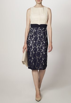 Abito blu navy e crema con sovrapposizione in pizzo, design senza maniche, corpetto plissettato e cintura in tessuto. Gli accessori includono una clutch crema.