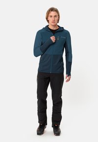 Giacca zip-up da uomo in verde scuro con parte superiore texturizzata. Presenta un cappuccio, una tasca con zip ed è abbinata a pantaloni neri e stivali.