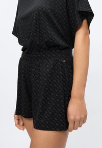 Γυναίκα που φοράει μαύρο σορτς ψηλής μέσης με διακριτικό γεωμετρικό μοτίβο και αντίστοιχο μαύρο αέρινο τοπ, σε απλό φόντο.