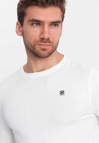 Witte lange mouwen t-shirt gemaakt van een gladde stof, met een klein zwart logo op de linkerkant van de borst. Ronde halsontwerp zonder extra patronen.