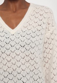 Pull blanc en tricot avec un motif en dentelle ajourée détaillé et un col en V, porté par une personne à la peau foncée.