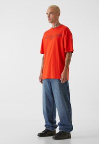 Oransje kortermet t-skjorte med svart tekst "REVELATION 12:07:12", kombinert med løse blå jeans og svarte joggesko.