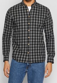 Camicia a quadri nera e grigia con colletto button-down e bottoni in legno, realizzata in tessuto testurizzato, indossata sopra jeans blu.