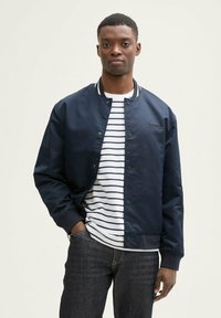 Chaqueta bomber azul marino con botones a presión, camiseta a rayas blanca y azul por debajo, y jeans oscuros. Tejido suave y detalles acanalados.