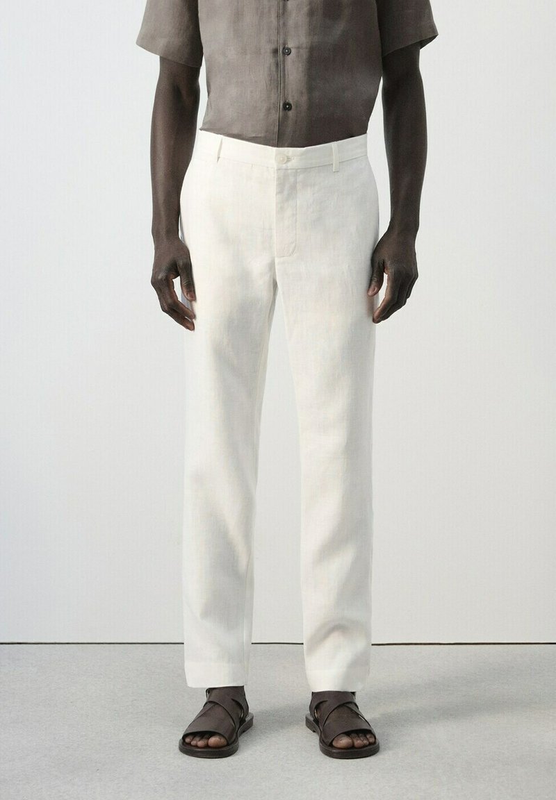 Man draagt witte rechte broek, een loshangend kortbruin overhemd met korte mouwen en donkerbruine sandalen met open tenen, staand op een grijze vloer.