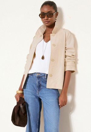 Frau trägt eine beige Jacke mit Knöpfen über einem weißen Oberteil, blaue Jeans, dunkle Sonnenbrille, goldene Creolen, eine Anhänger-Halskette und hält eine braune Tasche.