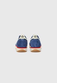 Blue suede sneakers with a white and red stripe on the sole, featuring a vintage-style logo on the heel and textured detailing.