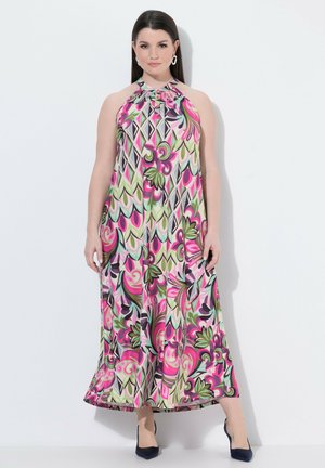 Frau mit langen dunklen Haaren, die ein ärmelloses Neckholderkleid mit einem bunten abstrakten Blumenmuster in Pink, Grün und Schwarz sowie schwarze Schuhe mit Absatz trägt.