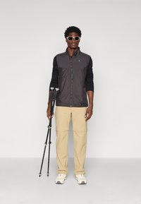 Chaleco ligero negro con cierre frontal, combinado con pantalones beiges desmontables y zapatillas blancas. La figura sostiene bastones de senderismo negros.