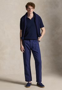 Młody mężczyzna stoi, ubrany w granatową koszulkę polo, dopasowane spodnie, sweter narzucony na ramiona, niebieskie espadryle i zegarek na nadgarstku, na neutralnym tle.