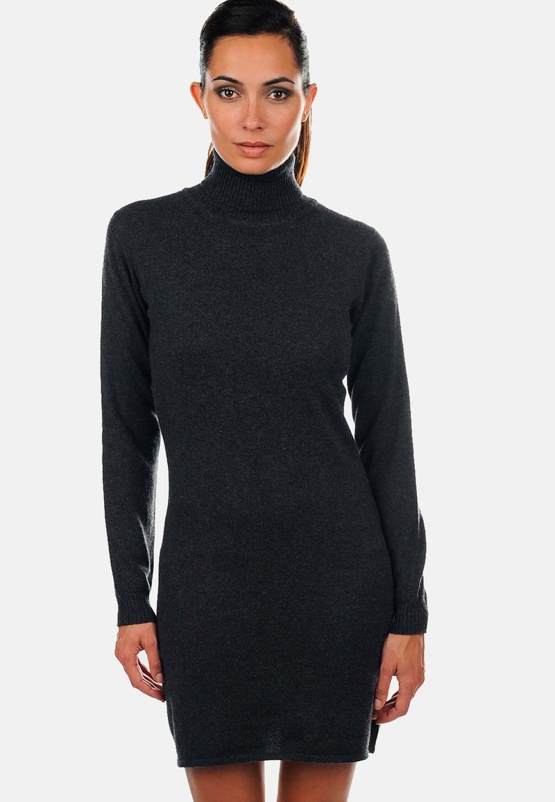 Femme portant une robe pull à col roule long en gris foncé, se tenant devant un fond blanc uni.