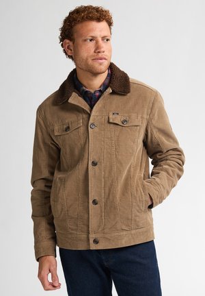 Man wearing a tan corduroy jacket with dark brown sherpa collar over a blue and red plaid shirt, posing with one hand in pocket.