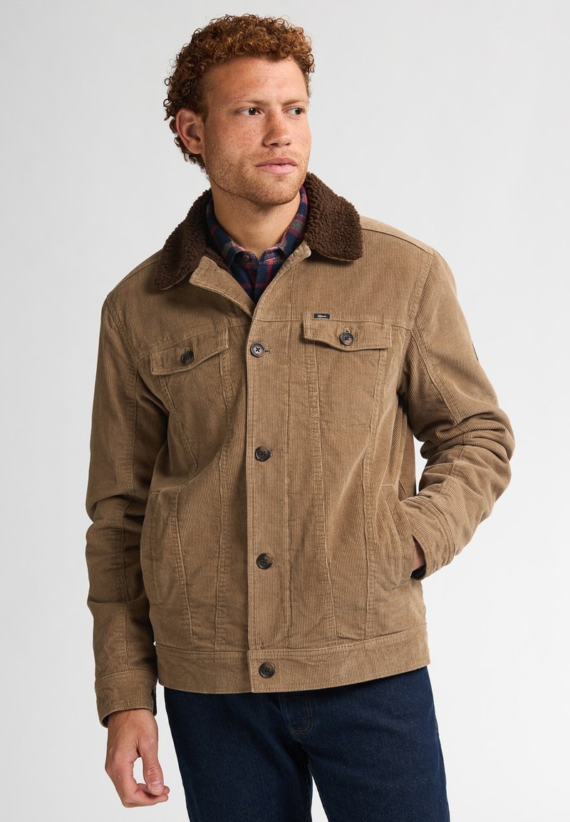 Uomo che indossa una giacca di velluto a coste color cammello con collare in sherpa marrone scuro sopra una camicia a quadri blu e rossa, posando con una mano in tasca.