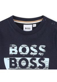 Kék pamut póló, amelyen a "BOSS" szó grafikája látható, elnyűtt világoskék színben, lekerekített nyakkal és patent gombokkal.
