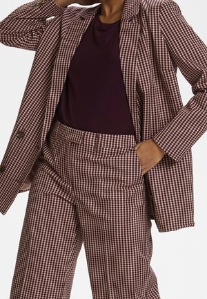 Person iført en brun- og hvidternede dragt med højtaljede bukser og matchende blazer over en mørkelilla skjorte, hånd i lommen.