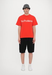 Rotes Baumwoll-T-Shirt mit dem Text "No Problemo", schwarze Shorts und weiße Sneakers. Model mit einer dunkelgrauen Mütze, steht vor einem weißen Hintergrund.