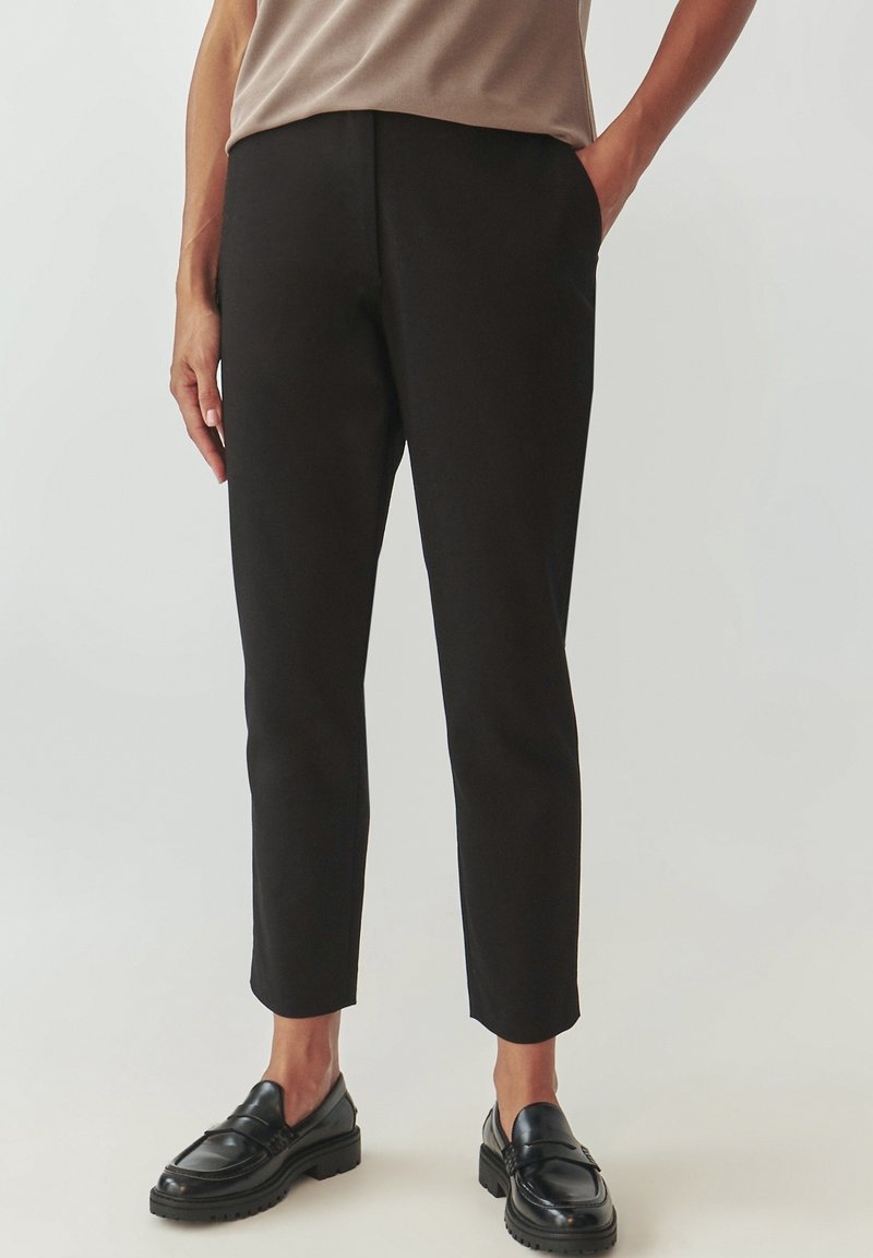 Personne portant un pantalon noir slim longueur cheville, des mocassins noirs et un haut beige, une main dans la poche, sur un fond uni.