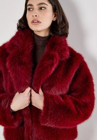 Burgunderfarbener Kunstpelzmantel mit einem breiten Kragen und lockerer Passform. Die weiche Textur, langen Ärmel und tiefen Taschen schaffen eine gemütliche Silhouette.
