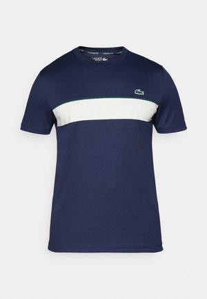 Camiseta de manga corta azul marino con una franja horizontal blanca y acentos verdes. Presenta el logotipo de Lacoste en el lado izquierdo del pecho. Textura suave.