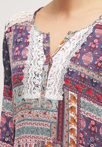 Blusa da donna multicolore con motivi floreali e geometrici, con bordino in pizzo bianco lungo il collo e la coulisse dei bottoni.