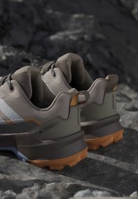Ζευγάρι μπεζ και γκρι παπούτσια trail running Adidas Terrex με πορτοκαλί σόλες σε βραχώδες έδαφος.