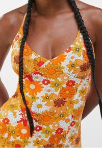 Nainen, jolla on tummat letitetyt hiukset, pukeutuu ilmanhihaiseen mekkoon, jossa on oranssia, keltaista ja valkoista kukkakuviota.