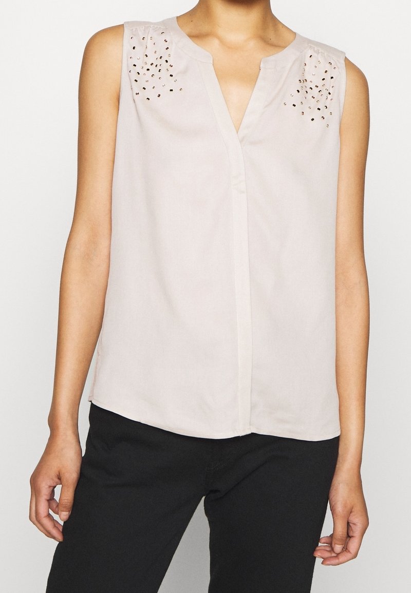 Blouse beige sans manches avec un col en V, ornée de clous dorés décoratifs sur les épaules. Tissu léger avec une texture lisse.