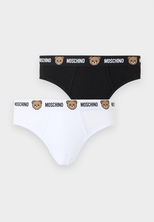 Izmos férfi mezítláb áll, fekete alsónadrágot visel, amelyen medvearc-minta és "Moschino" logó található a deréknál, egyszínű fehér háttér előtt.