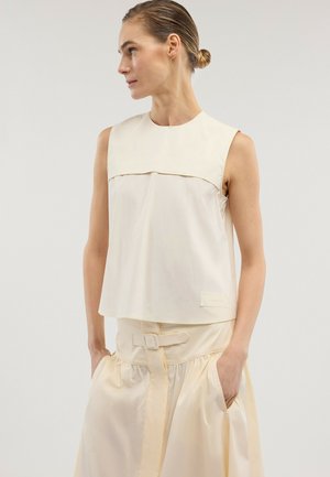 Frau mit zurückgekämmtem Haar, die ein ärmelloses cremefarbenes Oberteil und einen passenden, hoch taillierten Rock mit Gürtel trägt, die Hände in den Taschen, schaut seitlich.