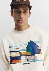 Crèmewitte sweatshirt met een grafische print van een landschapsbeeld in blauw- en groentinten en een geribde hals. Tekst: "207-213 STUDIOS."