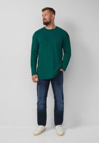 Groene lange mouwen shirt gemaakt van zachte stof, gecombineerd met donkerdenim jeans en lichte sneakers, staand tegen een neutrale achtergrond.