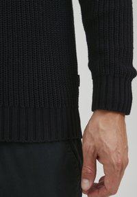 Navyblauer gestrickter Pullover mit geripptem Saum und Bündchen sowie einem strukturierten Muster. Etikett in der Nähe des unteren Randes sichtbar. Teilweise Hand gezeigt.