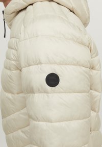 Giacca imbottita color beige chiaro con design trapuntato. Presenta una patch rotonda nera con logo sul lato sinistro. Materiale testurizzato e isolante.