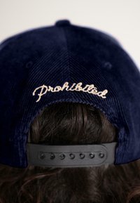 Rear view of a person wearing a navy blue corduroy baseball cap with adjustable strap and "Prohibited" embroidered in beige.
