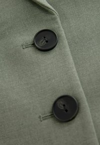 Deux boutons noirs attachés à travers des boutonnières cousues sur un tissu vert olive clair, probablement une partie d'un manteau ou d'une veste.