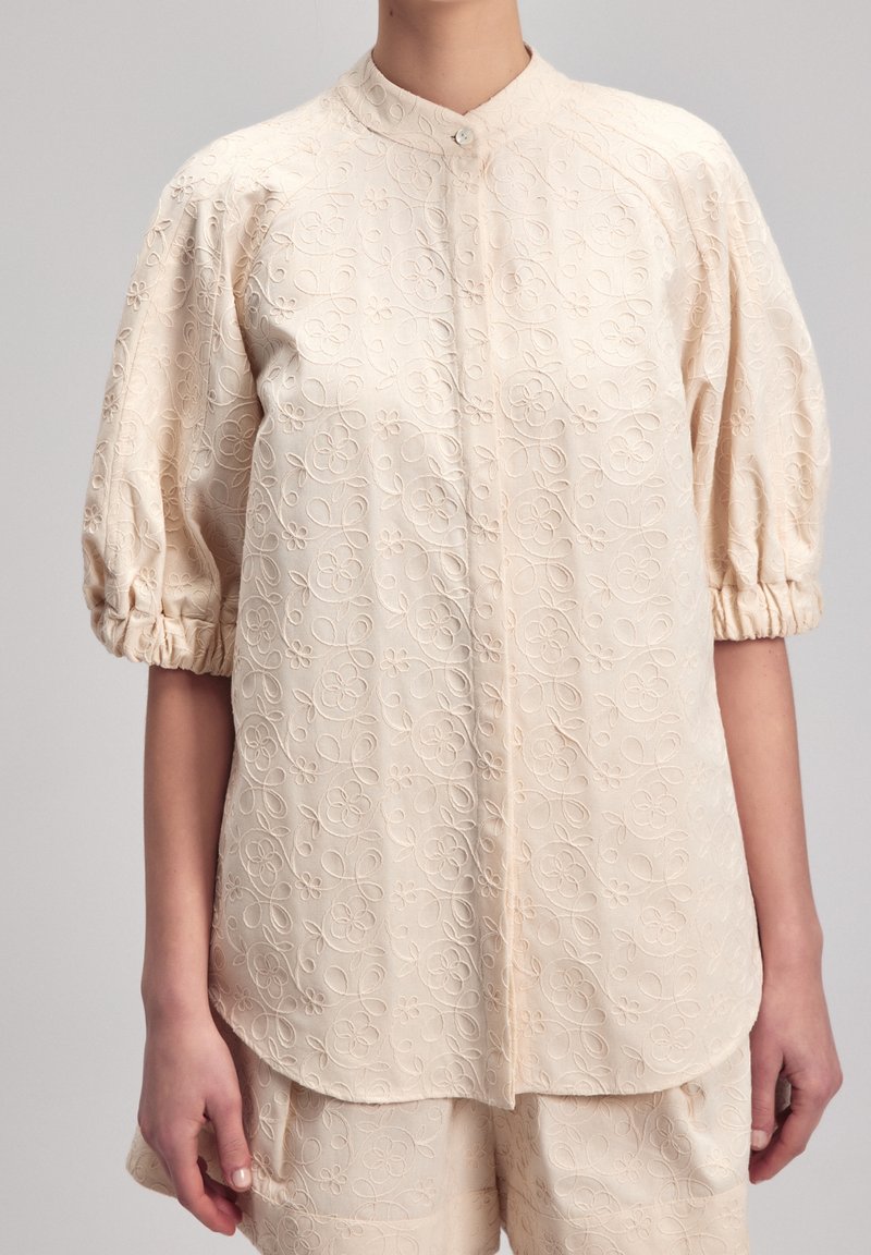 Blouse crème avec manches bouffantes aux coudes et broderie florale portée avec un short assorti sur fond uni.