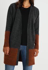 Lång kappa i mörkgrått och rost. Har ribbade ärmslut och nederkant, öppen design framtill och mjuk texturerad stickning. Kombineras med en svart topp.