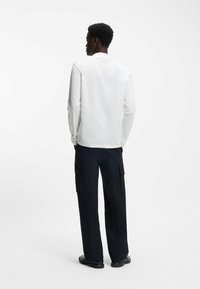 Weißes Langarmhemd und schwarze Cargo-Hosen mit Taschen, entspannter Passform und glatter Stoffoberfläche. Legere Schuhe runden den Look ab.