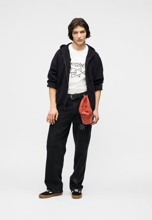 Tânăr purtând hanorac negru, tricou alb cu grafică, blugi negri largi cu ornamente, teniși negri, ținând o șapcă roșie pe lângă el.