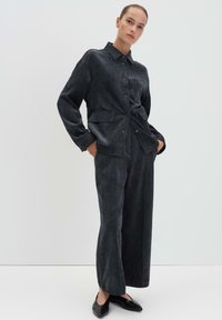 Completo in tessuto grigio scuro composto da una camicia con colletto e bottoni e pantaloni a gamba larga. Tessuto liscio con due tasche frontali.