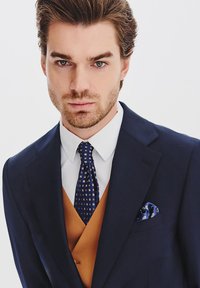 Granatowa marynarka na kremowej koszuli, z wzorzystym niebieskim krawatem i złotym kamizelką. Wkładka do kieszeni dodaje niebieskiego, kwiatowego akcentu.
