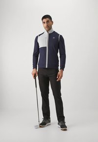 Tamsiai mėlyna ir balta užtrauktuku užsegama golfo striukė su aukštu apykakliu, derinama su juodomis kelnėmis ir pilkais golfo batais, laikant golfo lazdą.