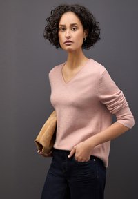 Suéter de color rosa claro, con escote en V y hecho de un material suave, que presenta puños acanalados. Combinado con jeans oscuros, sostiene un bolso color beige. El fondo es gris.