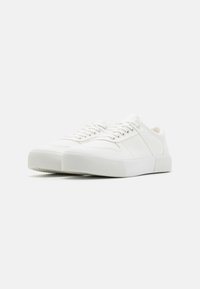 White canvas sneakers with rounded toe, flat laces, textured sole, and minimalistic design. Features subtle stitching and a clean aesthetic.