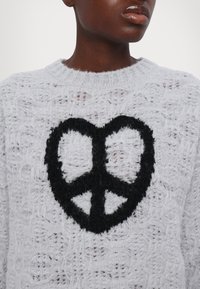 Γκρι πλεκτό πουλόβερ με χαλαρή πλέξη, που διαθέτει ένα έντονο μαύρο σύμβολο ειρήνης στο κέντρο. Μαλακή υφή με ορατές ίνες.