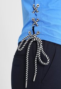 Niebieska koszulka z detalami sznurowania po bokach, z metalowymi oczkami w kolorze srebrnym i czarno-białymi sznurkami. Zbliżenie na materiał i teksturę.