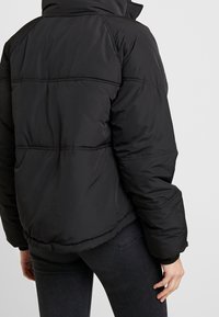 Černá péřová bunda s vysokým límcem, prošívaným designem a elastickými manžetami. Má mírně zkrácenou siluetu a jemnou, hladkou texturu.