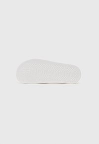 Semelle de chaussure en caoutchouc blanc présentant un motif en chevrons, avec un nom de marque en relief et une texture propre et lisse.