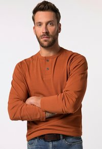 Långärmad skjorta i rostfärg med rund halsringning och knappslå med två knappar, tillsammans med blå denimjeans och ett brunt bälte.